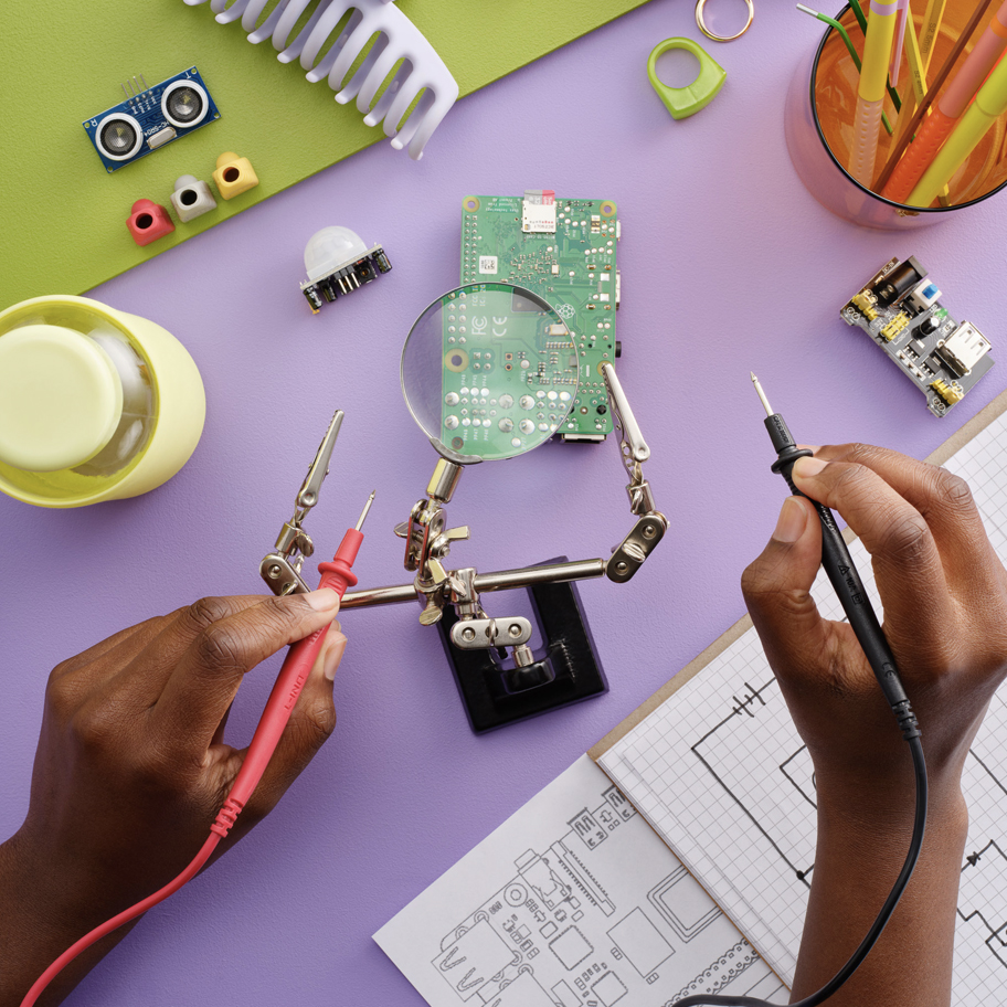 Bilden visar en arbetsyta för elektronik och teknikutveckling, där en person arbetar med en kretskort. Händerna håller två testprober, en röd och en svart, vilket tyder på att personen testar eller felsöker elektroniska komponenter. Kretskortet är fastspänt i en justerbar hållare med en förstoringsglas, vilket underlättar precisionsarbete. Runt arbetsytan finns olika elektronikdelar, som en ultraljudssensor, komponenter, ritningar av kretsar och anteckningar på rutat papper. Färgglada föremål, som pennor och en grön arbetsmatta, skapar en kreativ och inspirerande atmosfär.